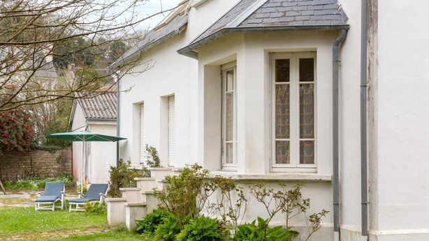 A white house with a bay window, a small garden with grass and plants, and two lounge chairs.