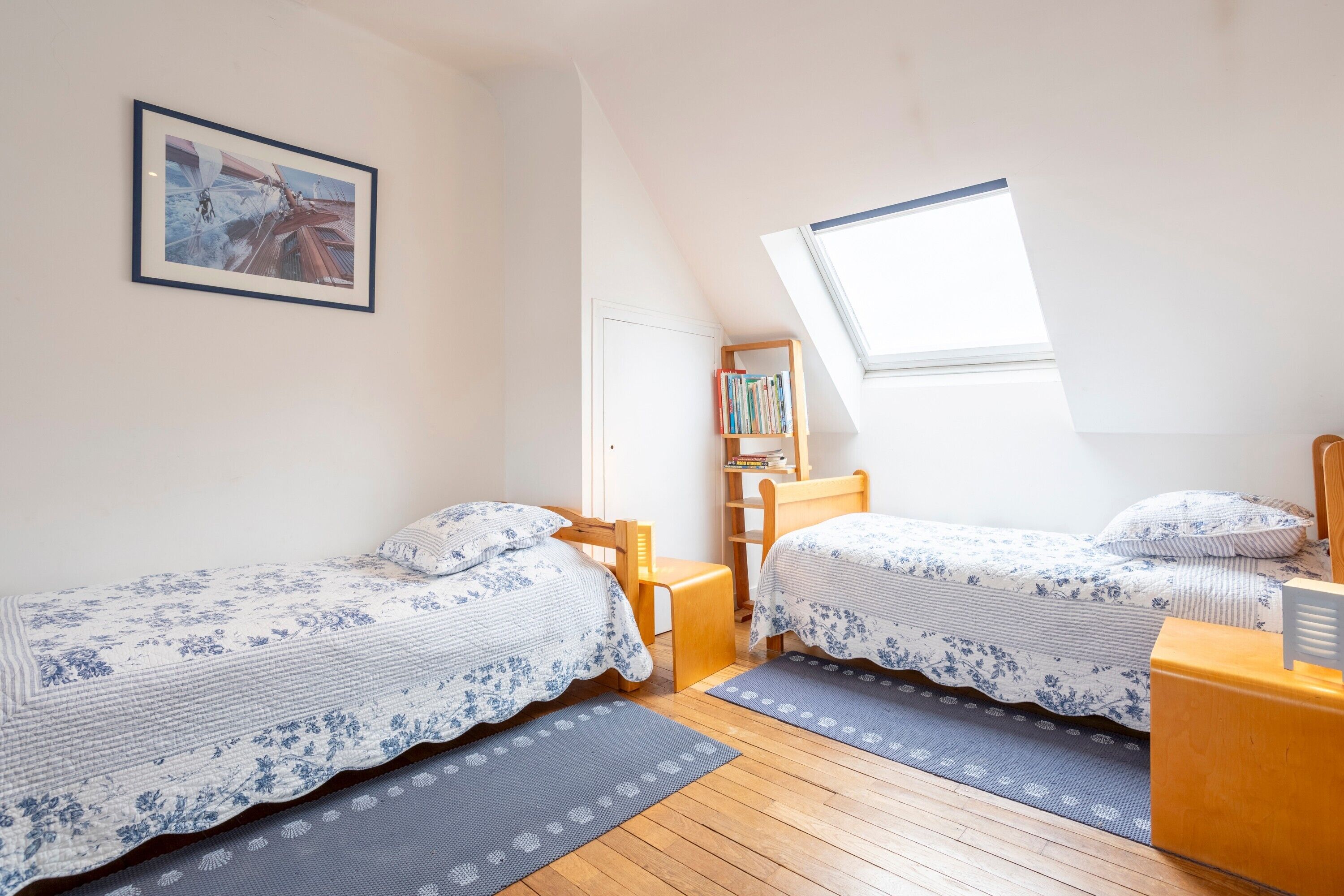 Een slaapkamer met twee bedden, een dakraam, een boekenplank en een ingelijst schilderij aan de muur.
