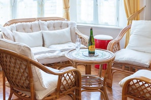 Un conjunto de sofá de mimbre con cojines, una mesa redonda con una botella y un vaso, y un taburete rojo.
