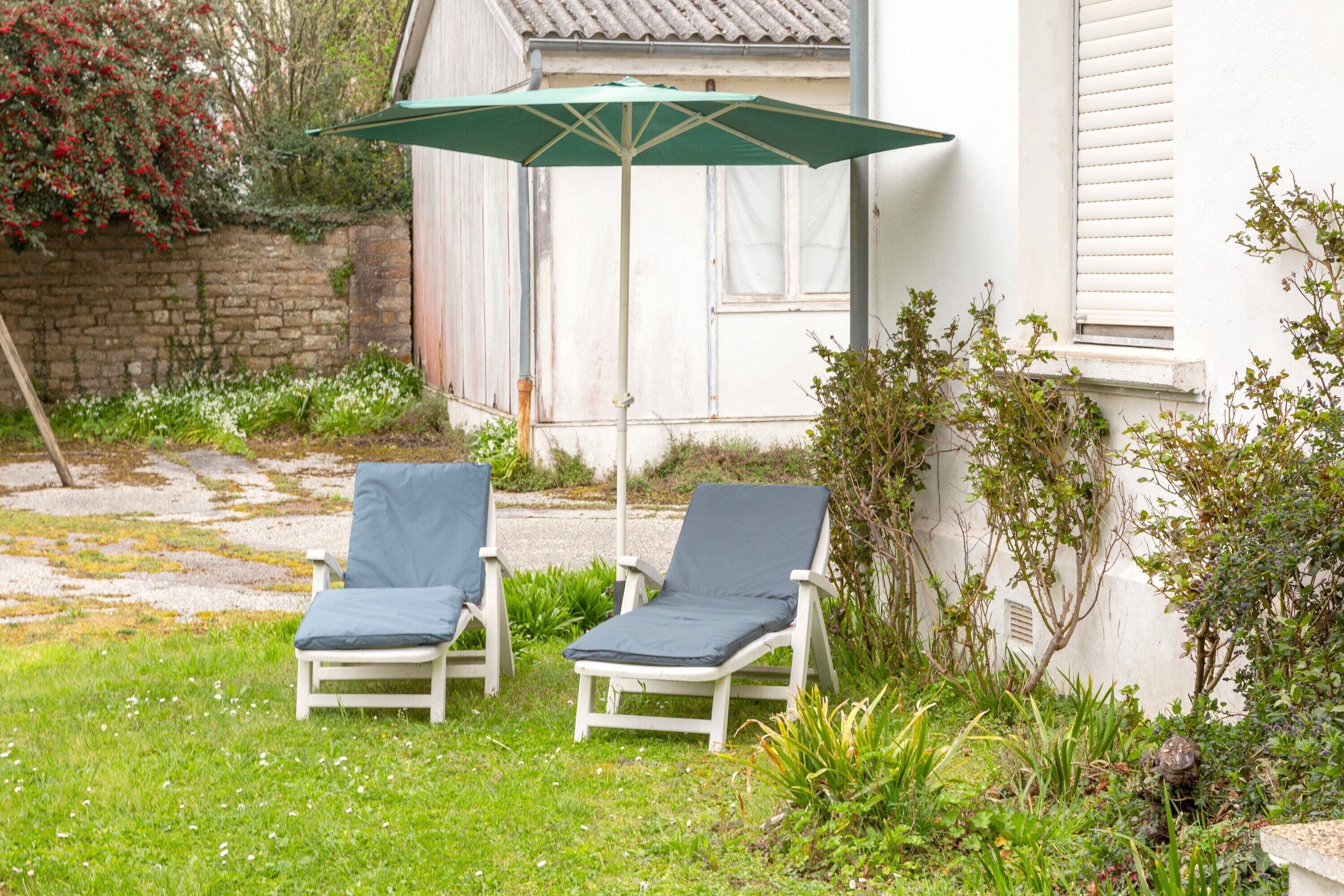 Dos sillones blancos con cojines azules, una sombrilla verde y una pared de piedra en un jardín.