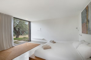 A modern bedroom with a large window, a bed with white linens, a bedside table, and a painting on the wall.