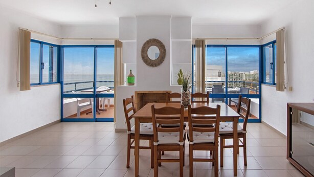 A dining area with a wooden table and chairs, a ceiling fan, a view through large windows, and a wall-mounted mirror. - Apartment 'T2 2 Linha Praia Vista Mar' with Sea View, Balcony and Wi-Fi (Unknown)