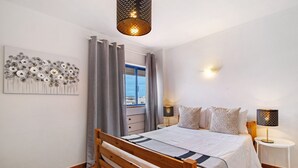 A bedroom with a wooden bed, a nightstand with a lamp, a wall art piece, and a view through a window with curtains. - Apartment 'T2 2 Linha Praia Vista Mar' with Sea View, Balcony and Wi-Fi (Unknown)