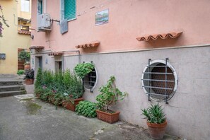 En bygning med terracotta tagsten, en lille have med plantede krukker, og et vindue med en metalgitter.