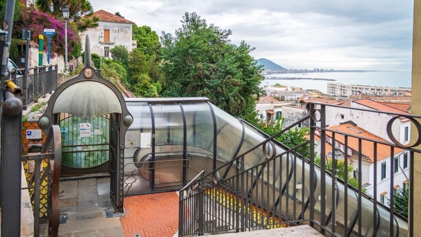 Un escalier avec une structure en verre menant à une terrasse offrant une vue sur la ville et la mer.