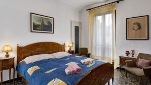 A hotel room with a bed, two bedside tables with lamps, a chair, a small table with decorative items, and a painting on the wall. - Holiday Home 'Dello Zio Birba' with Balcony and Wi-Fi (Portacomaro)