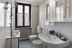 Une salle de bain avec un lavabo blanc, des toilettes, un bidet, une douche et une fenêtre avec des rideaux.
