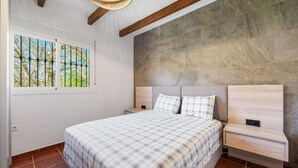 A bedroom with a large bed, two bedside tables, a headboard, and a window with a view of trees.