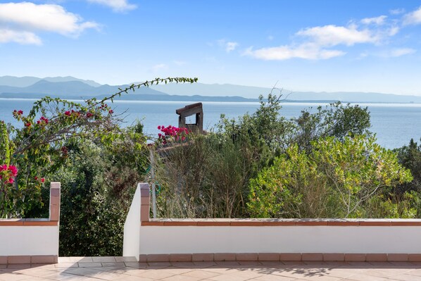 A terrace with a view of a lake and mountains, surrounded by greenery and a white wall.