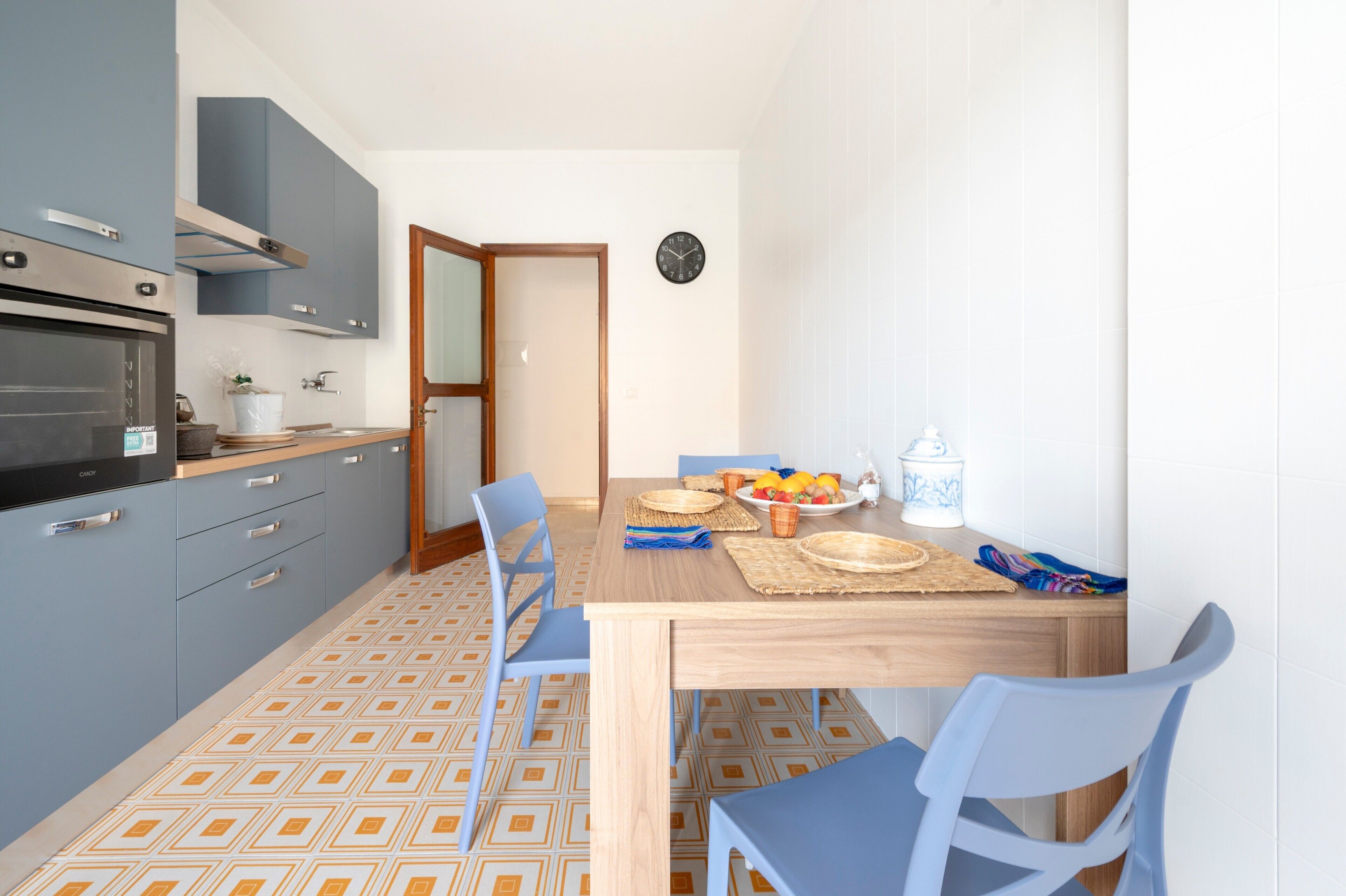 A compact kitchen with a built-in oven, a dining table with blue chairs, and a wall clock.