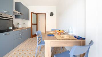 A compact kitchen with a built-in oven, a dining table with blue chairs, and a wall clock.
