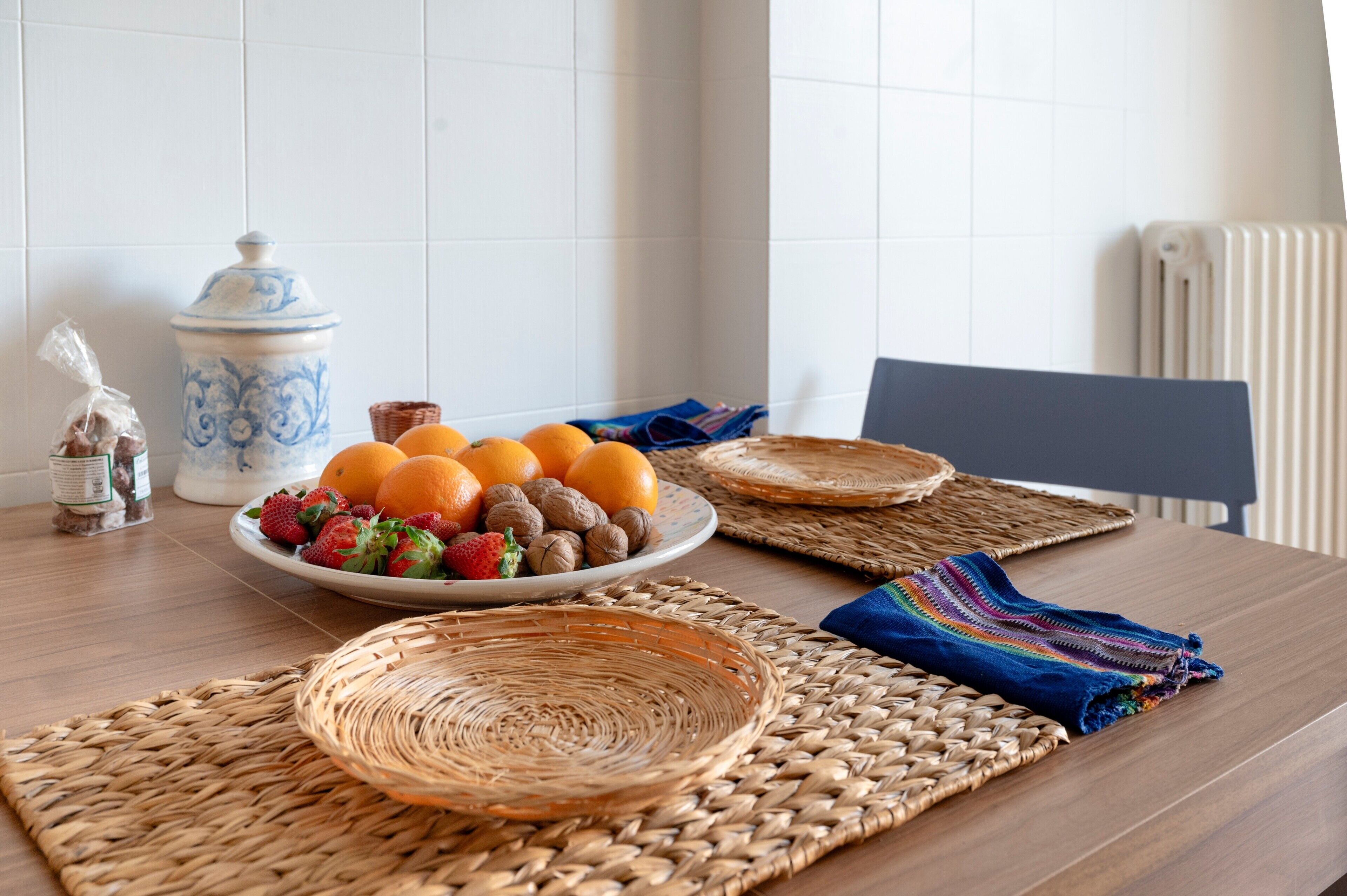 Un tavolo da pranzo con un piatto di frutta, una brocca di ceramica e tovagliette intrecciate.