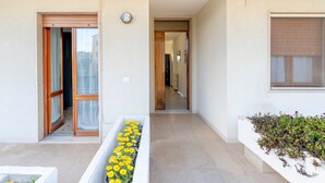 A building entrance with a sliding glass door, a concrete planter with yellow flowers, and a window with a view of buildings. - Apartment 'Stella' with Sea View, Wi-Fi and Air Conditioning (Otranto)