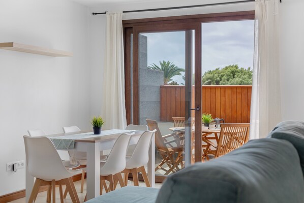A modern room with a dining area, a sofa, and a sliding glass door leading to an outdoor space with a wooden deck and a view of greenery.