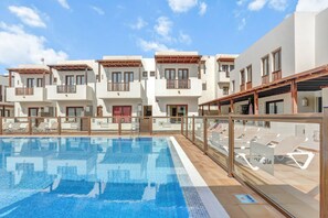A swimming pool with lounge chairs, a covered seating area, and a row of white buildings with balconies.