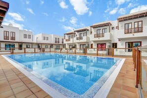 A swimming pool surrounded by modern buildings with balconies, a clear sky, and a tiled floor.
