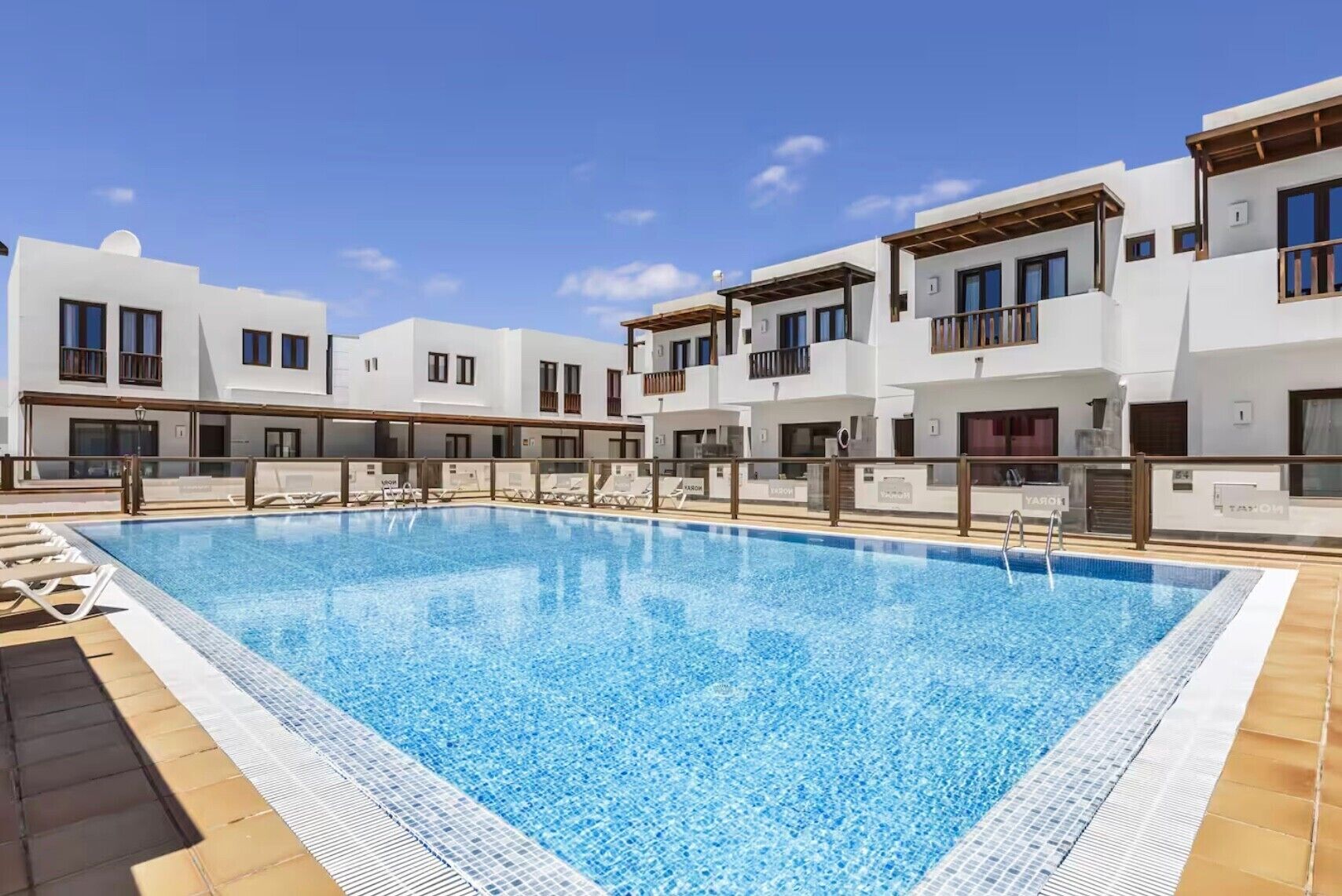 A swimming pool with lounge chairs, white buildings with balconies, and a clear sky.