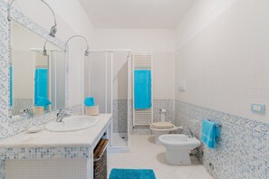 Un baño con un lavamanos blanco, un inodoro, un bidé, una ducha con puerta de cristal y un espejo con una toalla colgada.