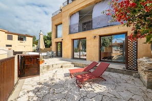 A two-story building with a patio area featuring stone flooring, a wooden gate, and red lounge chairs.