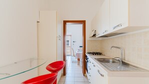 Une cuisine étroite avec des meubles blancs, un évier en acier inoxydable et une étagère en verre.
