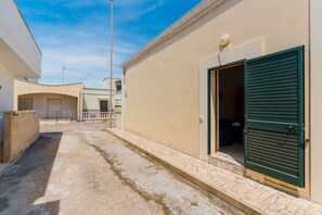 Une ruelle étroite, avec un bâtiment sur la droite, une porte à volets verts et un ciel dégagé.