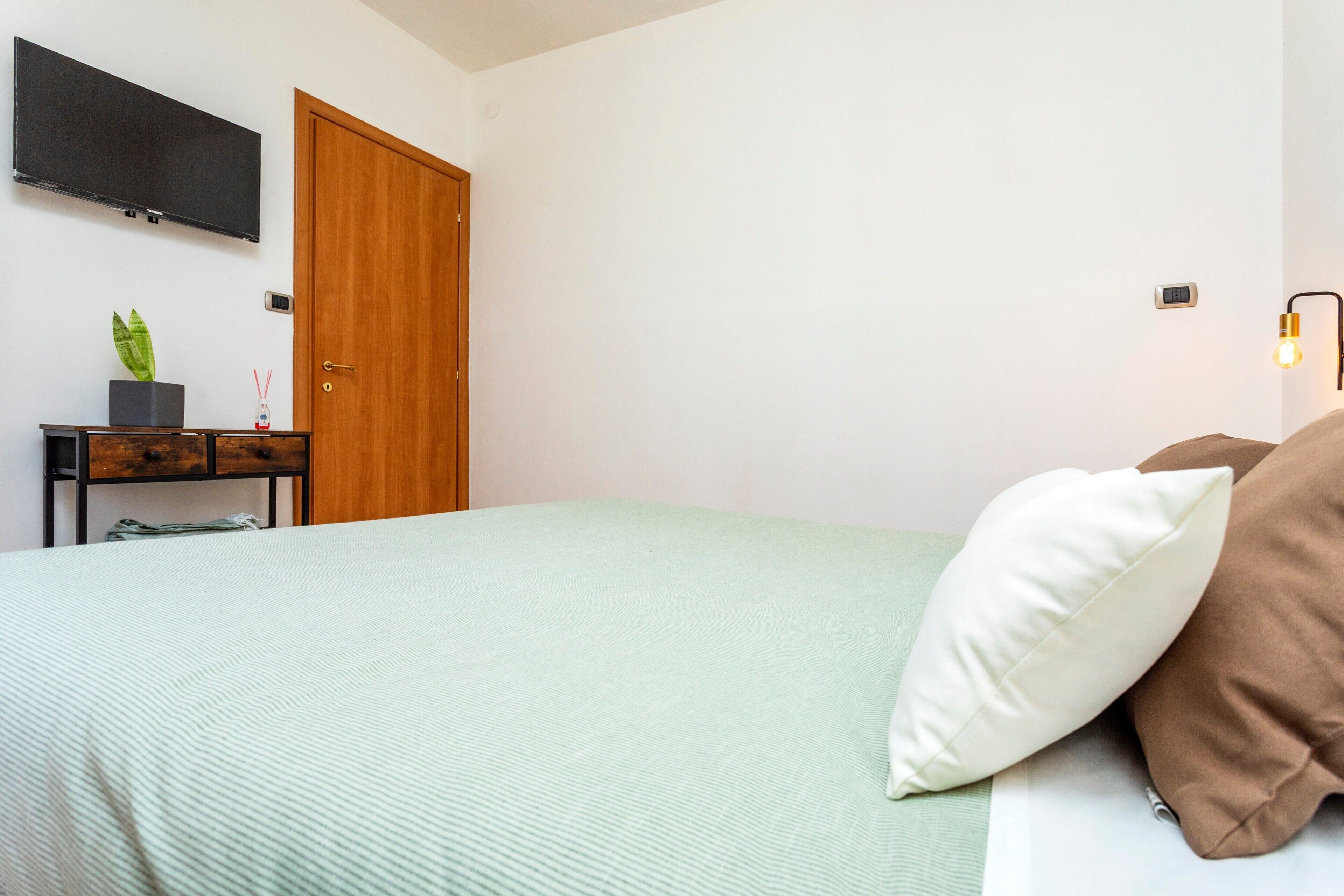 A bedroom with a bed, a TV mounted on the wall, a wooden door, a small table with a plant, and a wall-mounted light fixture.