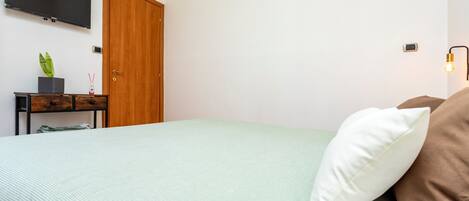 A bedroom with a bed, a TV mounted on the wall, a wooden door, a small table with a plant, and a wall-mounted light fixture.