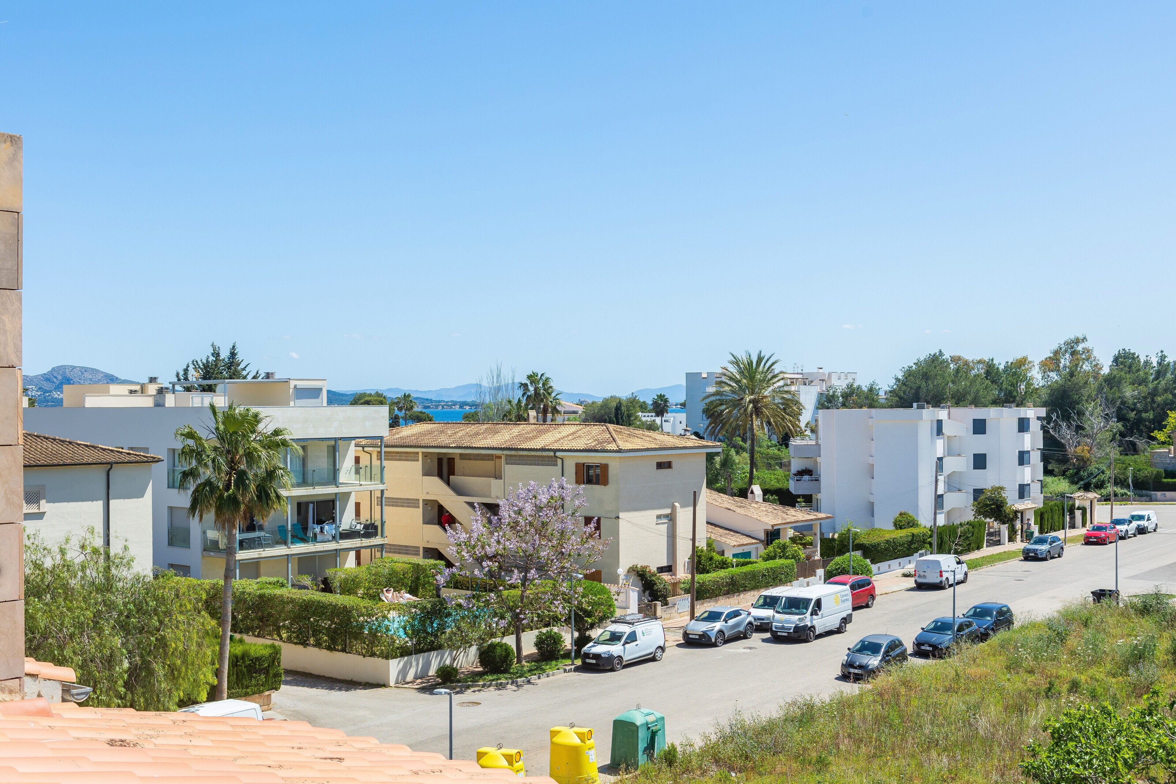 A residential street with parked cars, a building with balconies, and a clear blue sky.