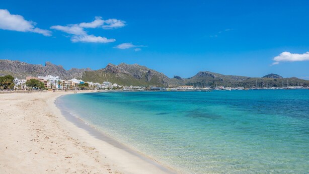A beach with clear turquoise water, a sandy shore, and a mountainous backdrop. - Apartment Planeta Mallorca in Puerto de Pollensa with Sea Views, Wi-Fi, and Air Conditioning (Unknown)
