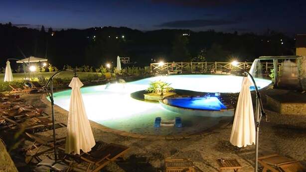 Zona de piscina nocturna con sillones de descanso y sombrillas, rodeada por una terraza de piedra.