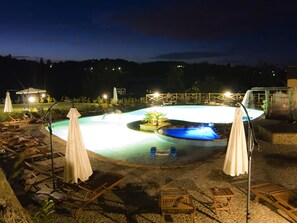 Area piscina notturna con poltrone a sdraio e ombrelloni, circondata da un solarium in pietra.