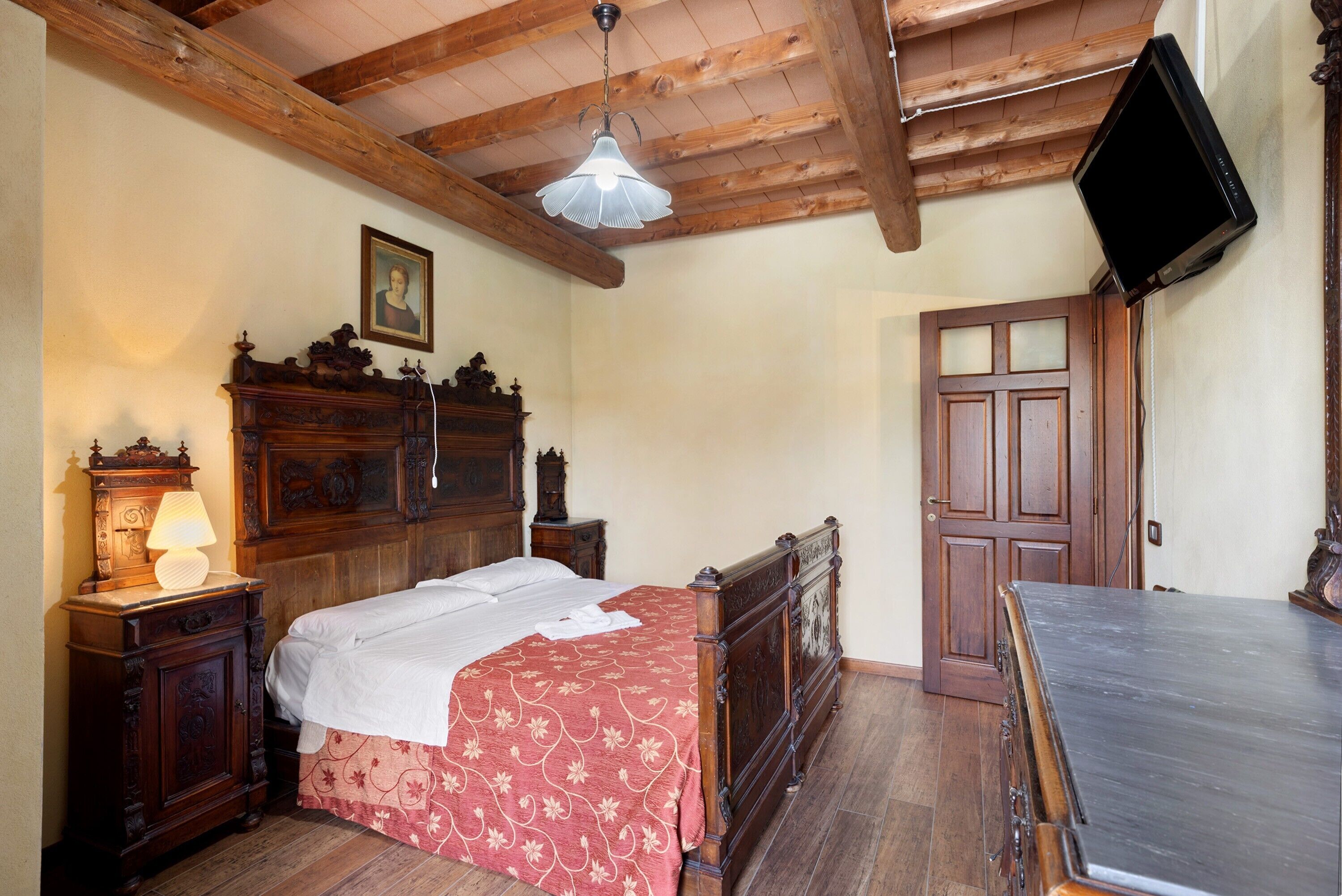 A bedroom with a wooden bed, a nightstand with a lamp, a dresser, a television mounted on the wall, and a wooden door.