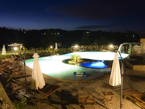 A nighttime pool area with lounge chairs and umbrellas, surrounded by a stone deck.