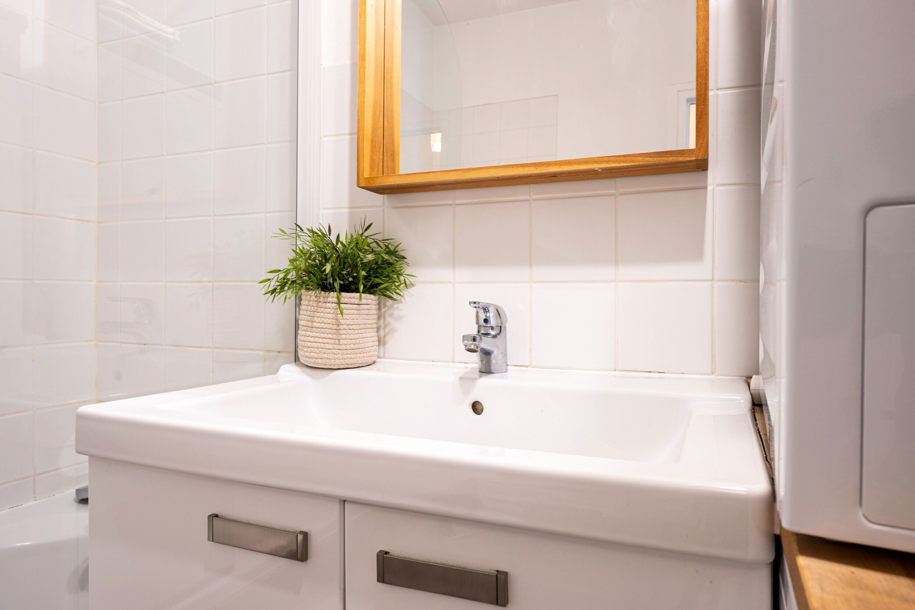 A bathroom with white tiles, a white sink with a wooden-framed mirror, a potted plant, and a wooden cabinet.