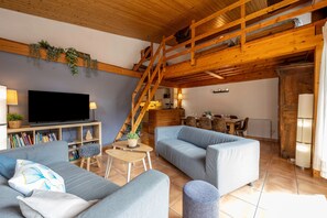 Una sala de estar acogedora con televisor, sofás, una escalera de madera y una cocina al fondo.
