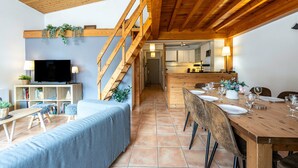 A modern interior with a wooden ceiling, a TV, a sofa, a dining table set for four, and a kitchen in the background.