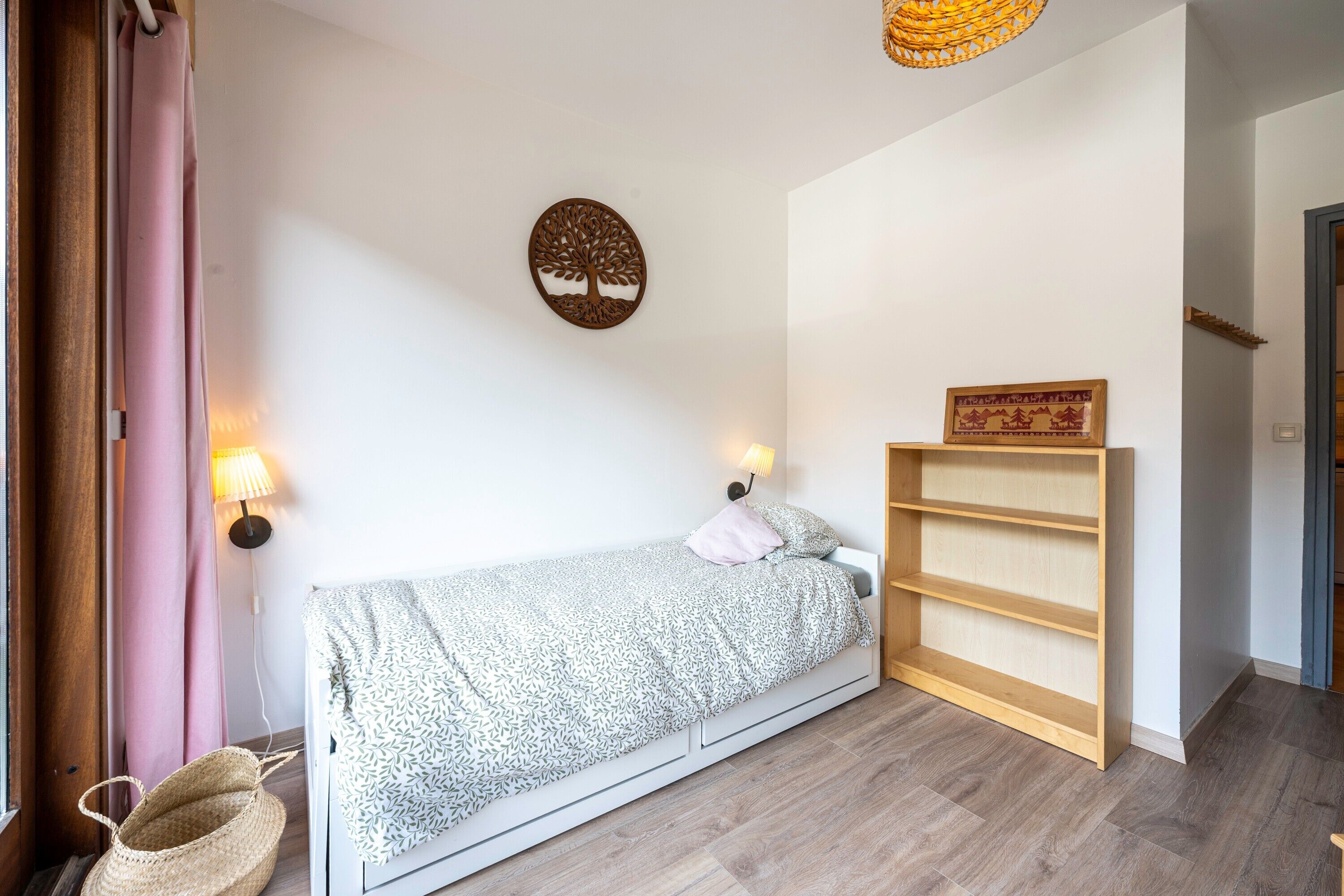 Un dormitorio pequeño y ordenado con cama, lámparas de noche, estante en la pared y una cesta tejida.