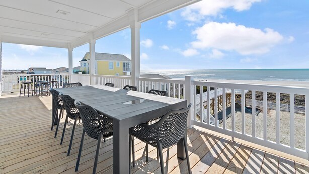 Una terraza cubierta al aire libre con una mesa y sillas, con vista a una playa y una casa.