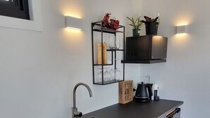 A modern kitchen with a dark countertop, a built-in sink, and a black cabinet. There are two wall-mounted light fixtures and a shelf with decorative items. - Tranquil Studio Retreat in Leura with Private Bush Outlook. (Leura)