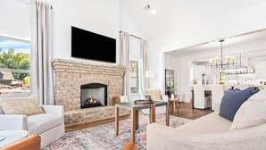 A modern living room with a fireplace, a flat-screen TV, and a dining area with a chandelier. - Plainsman Retreat-4min to AU, Gameroom,Pickleball+ (Auburn)