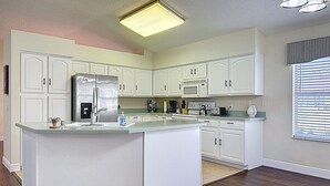 Une cuisine moderne avec des meubles blancs, un îlot central, des appareils en acier inoxydable et une grande fenêtre équipée de stores.