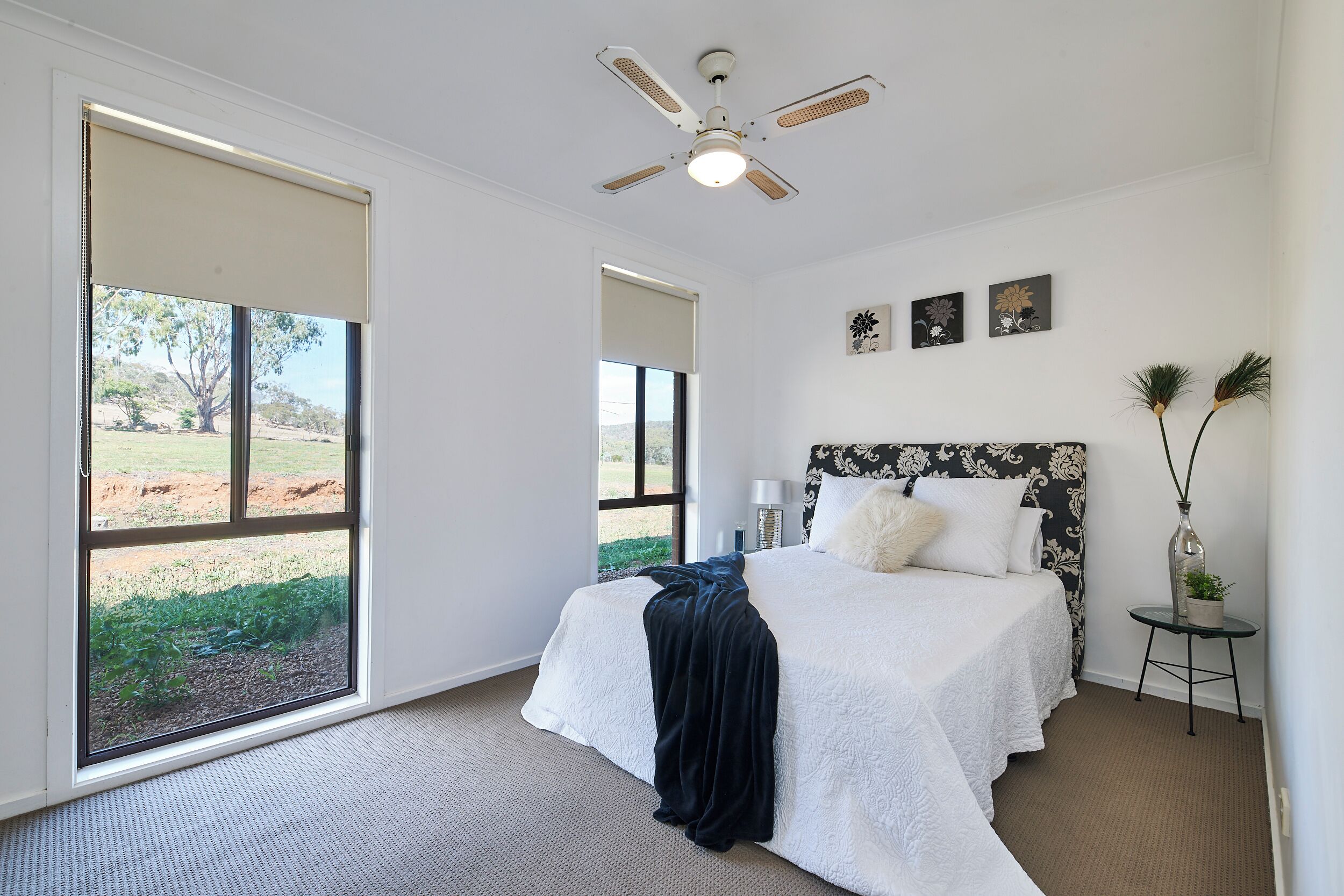 A bedroom with a large window, a ceiling fan, a bed with a white bedspread, and a small bedside table with a lamp and decorative items.