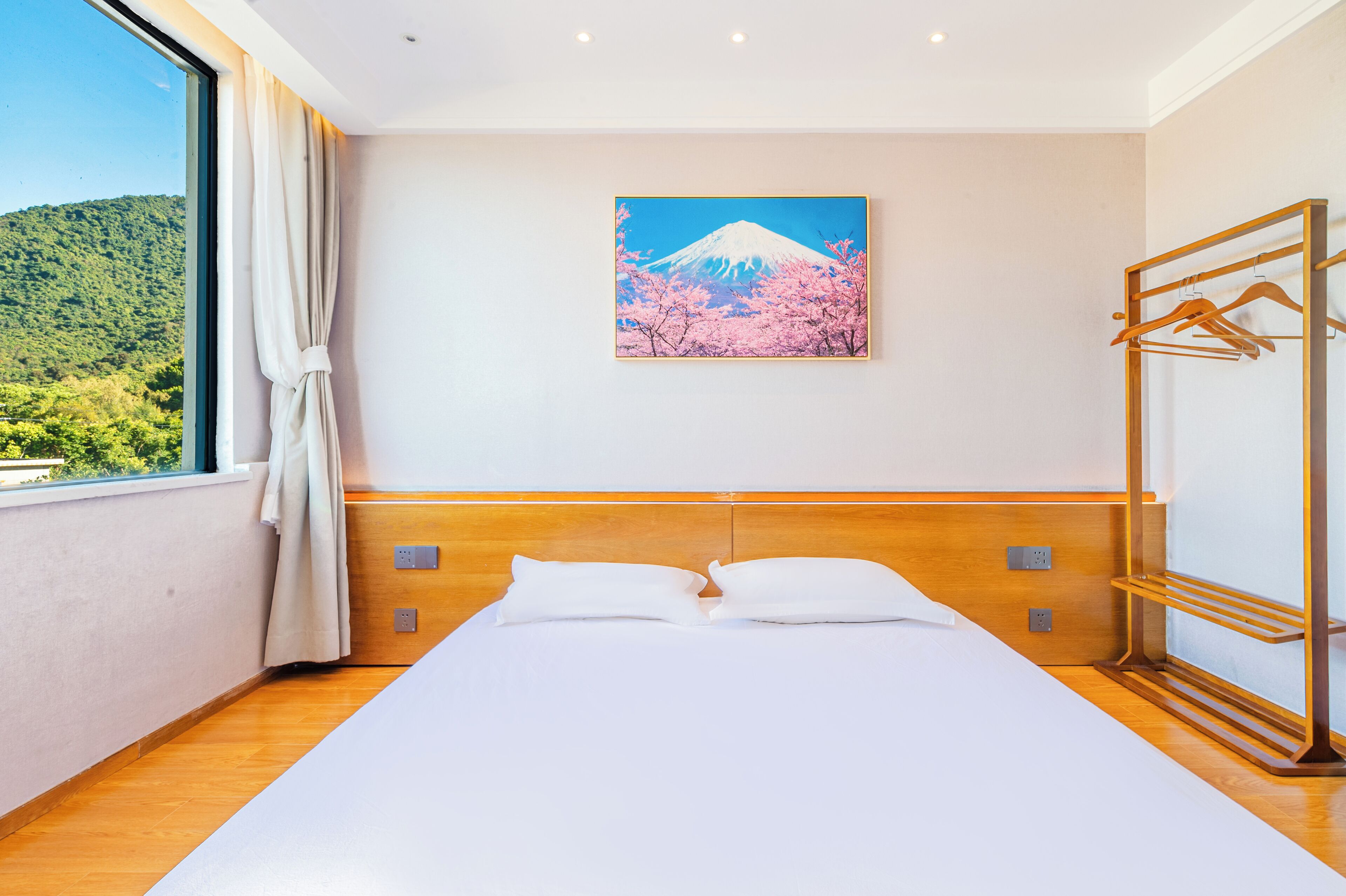 A hotel room with a bed, a wooden headboard, a window with a curtain, and a wall-mounted painting of a mountain and cherry blossoms.