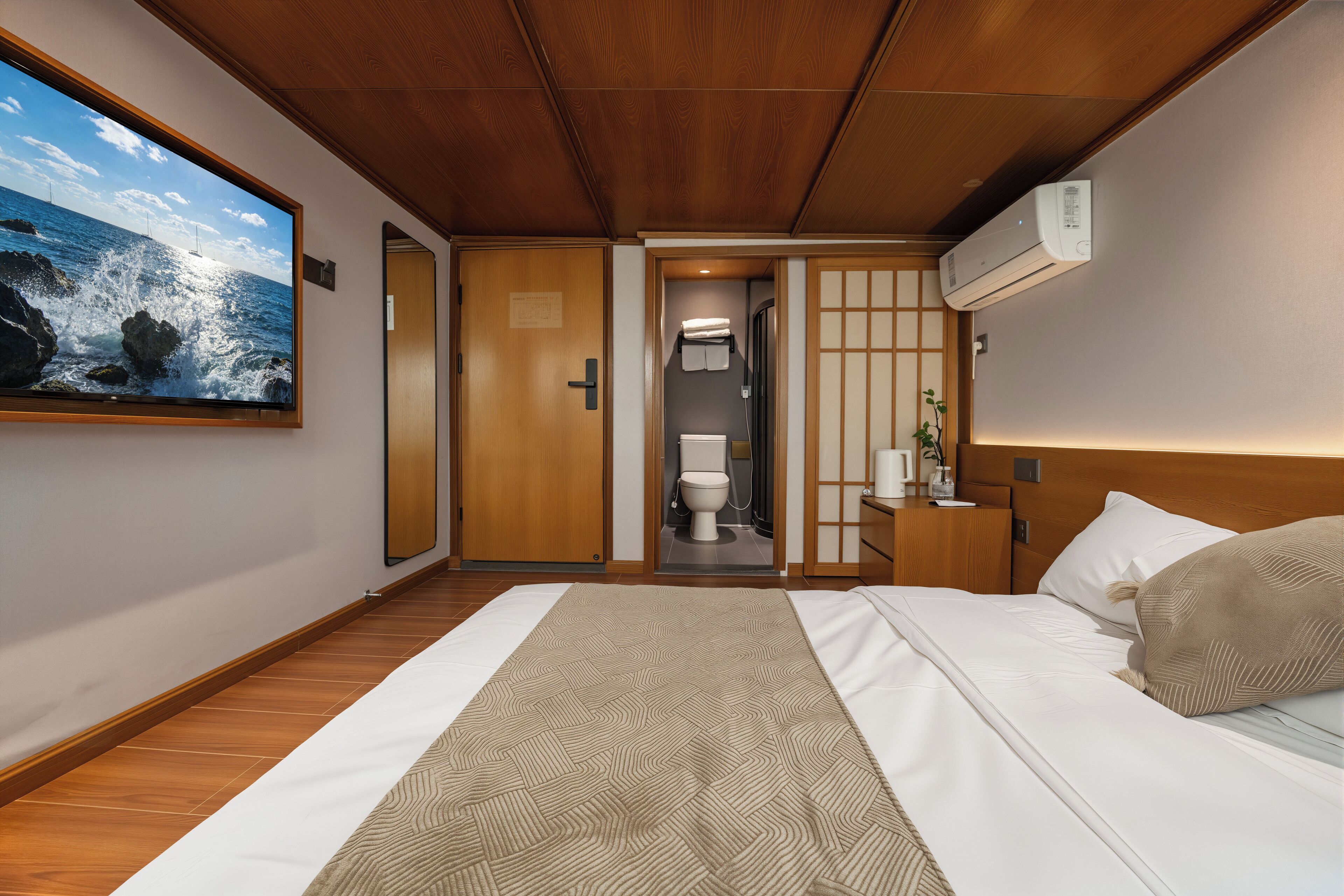A hotel room with a wooden ceiling, a large window with a view of the sea, a bed with white linens, a bedside table with a lamp, and a wall-mounted air conditioner.