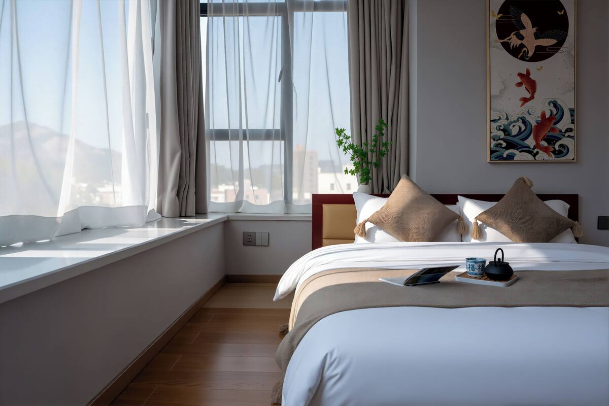 A bedroom with a large window, a bed with white bedding, a bedside table with a cup and a book, and a wall-mounted artwork.