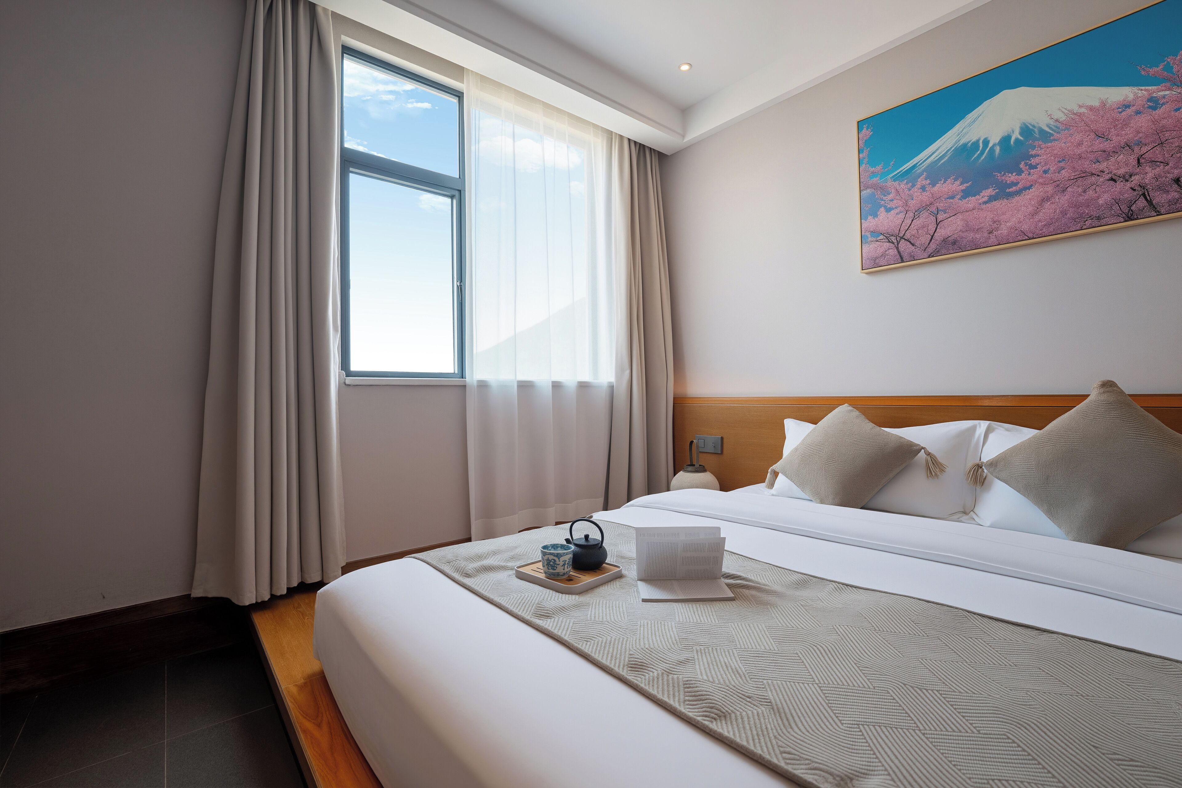 Ein Hotelzimmer mit einem großen Bett, einem Fenster mit Vorhängen, einem Gemälde vom Fuji und einem Nachttisch mit Wasserkocher und Tasse.