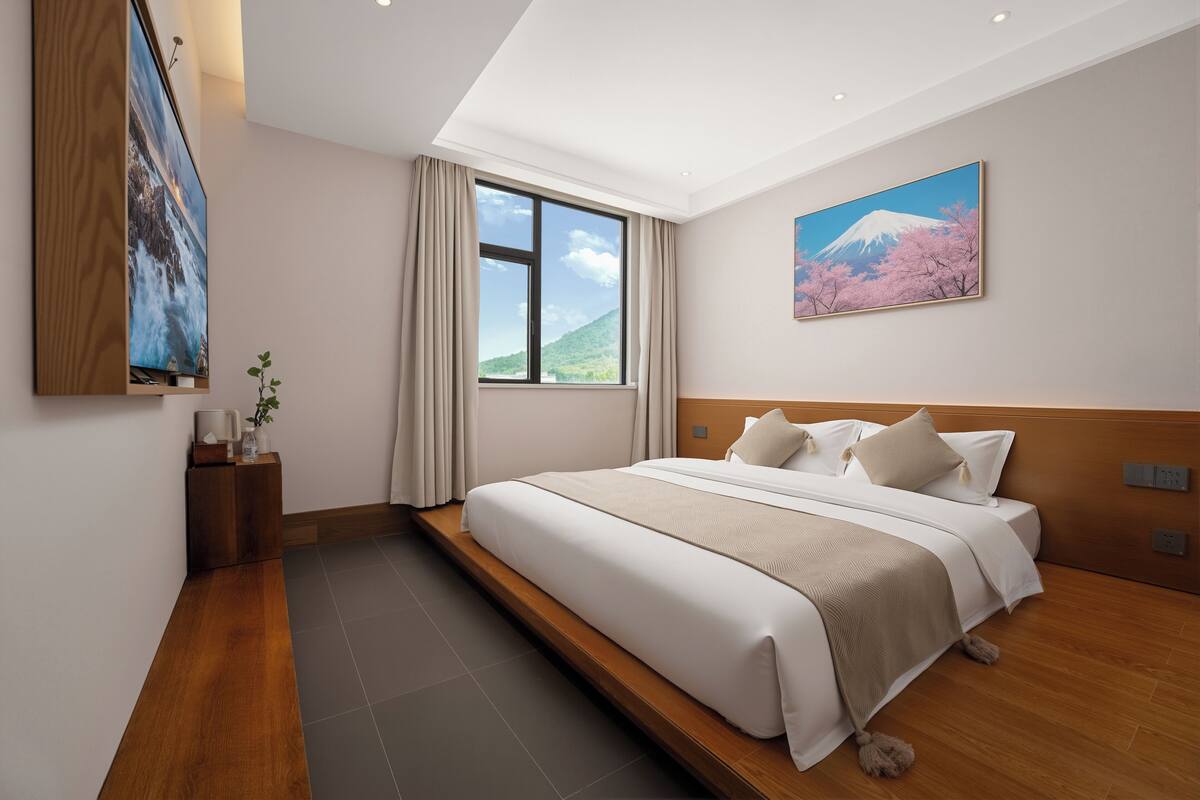 A modern hotel room with a large bed, a window with a view of a mountain, and a painting of cherry blossoms.