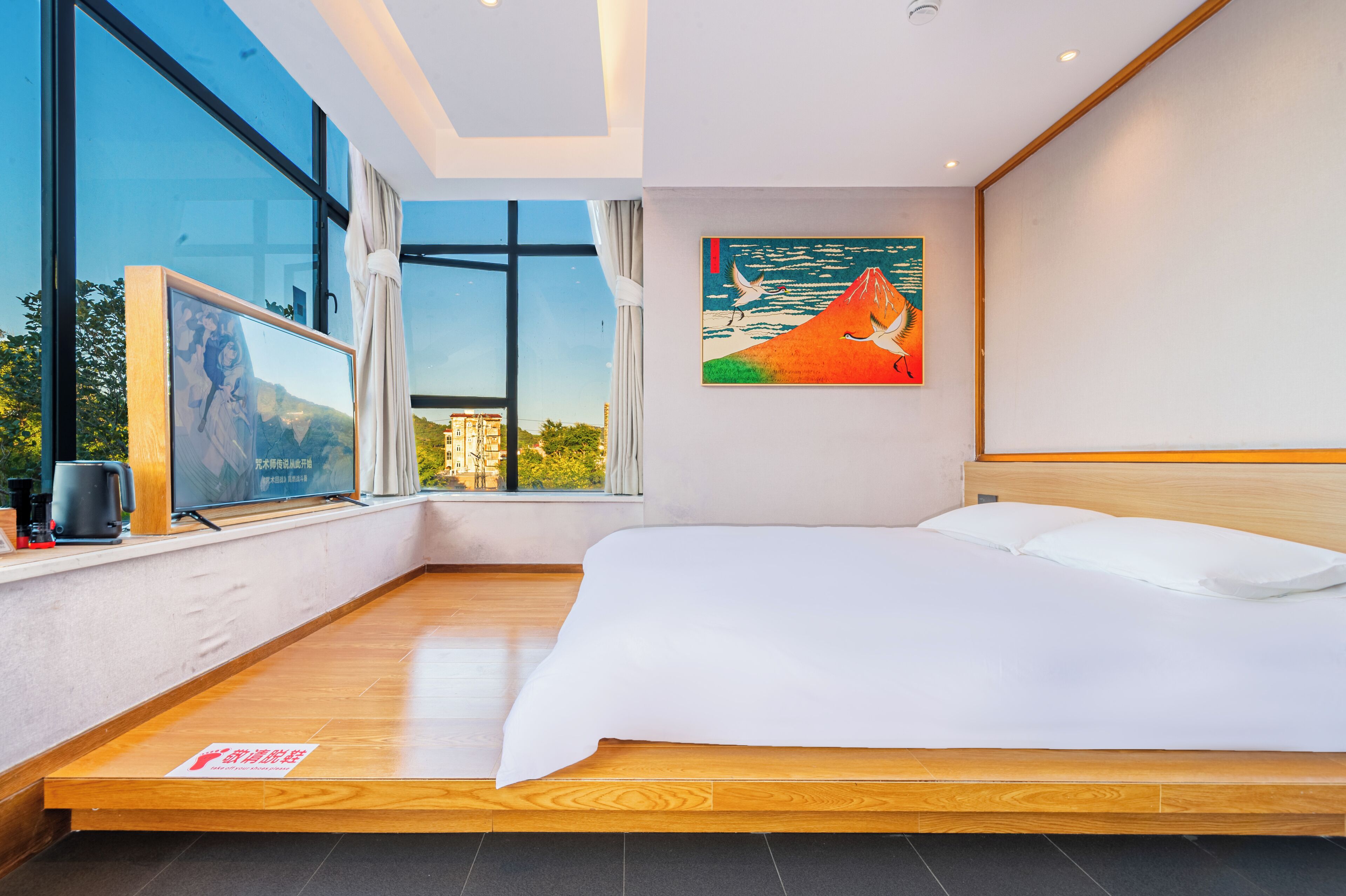 A modern hotel room with a large bed, a painting of a mountain and seagulls, a window with a view of buildings, and a wooden floor.