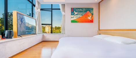 A modern hotel room with a large bed, a painting of a mountain and seagulls, a window with a view of buildings, and a wooden floor.