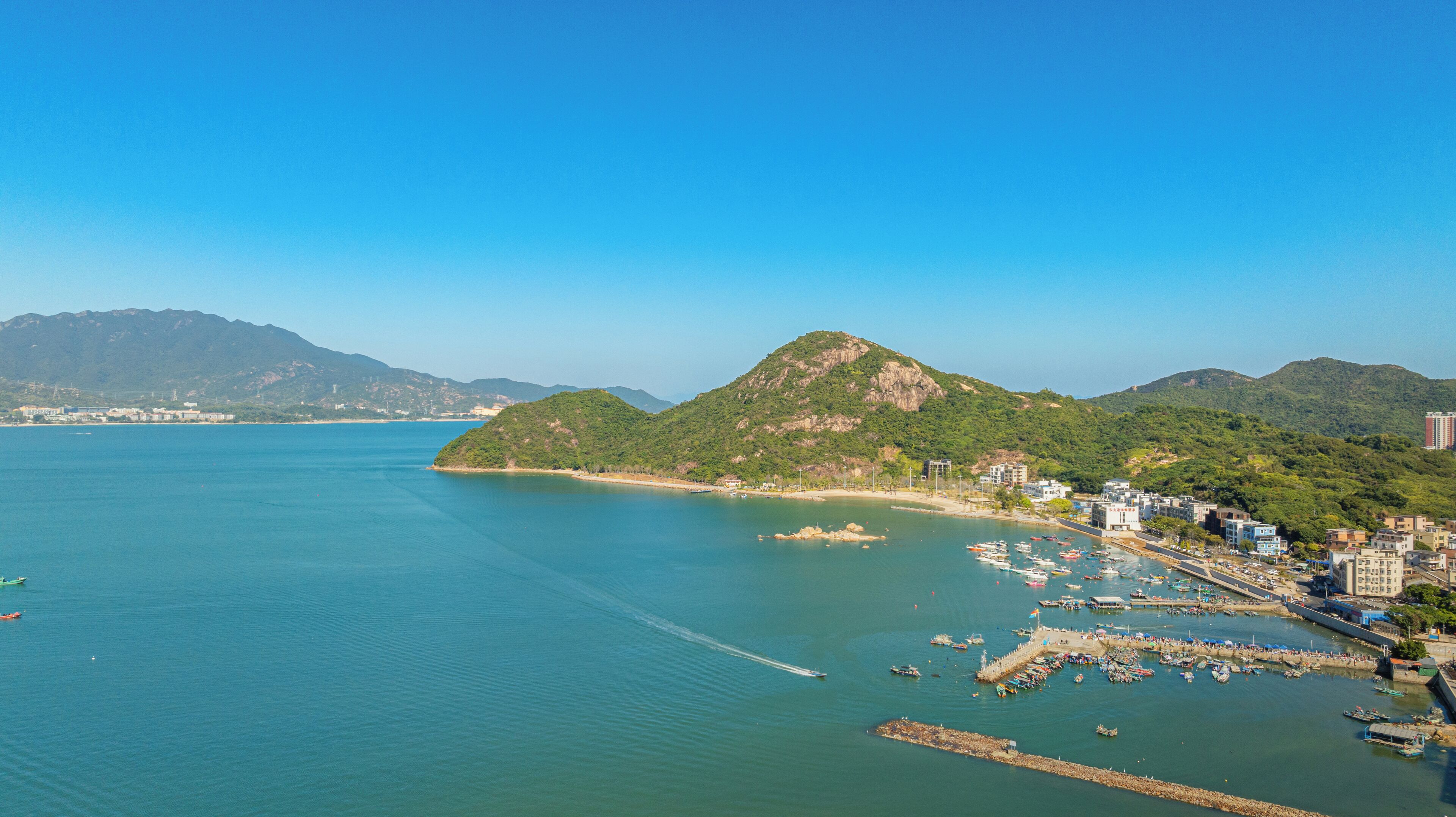 Uma cidade costeira com um porto de iates, edifícios ao longo da costa e uma colina imponente ao fundo.
